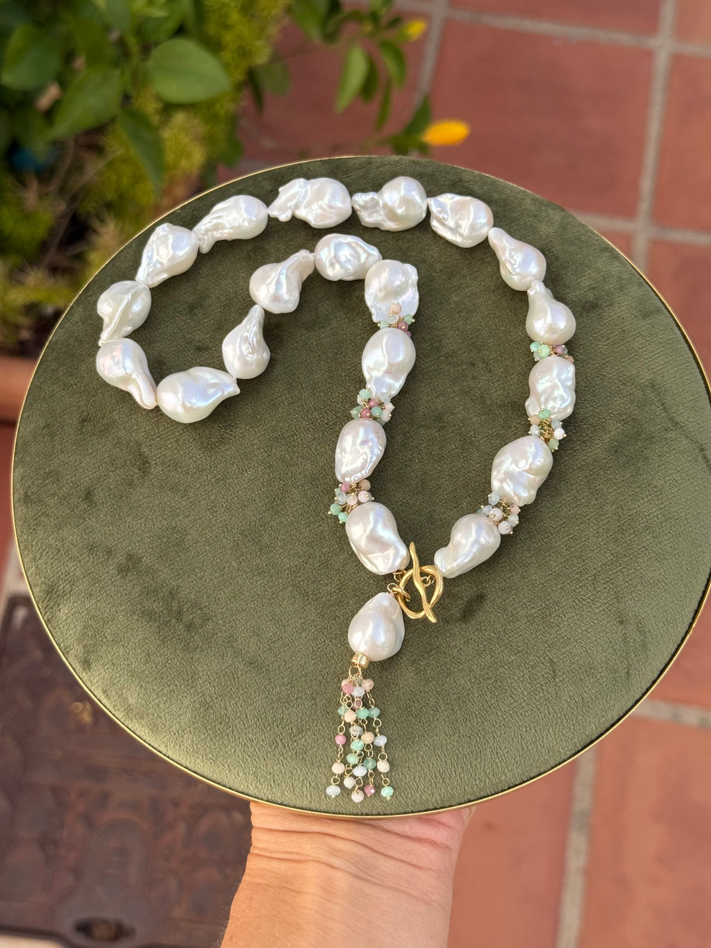 Fiora statement necklace with irregular white baroque pearls, gemstone accents and dangling pearl tassel on green display
